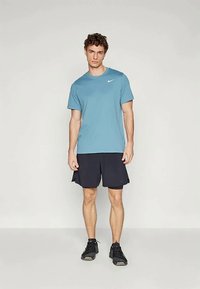 Ung mann som står iført en blå Nike T-skjorte, svarte treningsshorts og svarte joggesko mot en enkel lys bakgrunn.