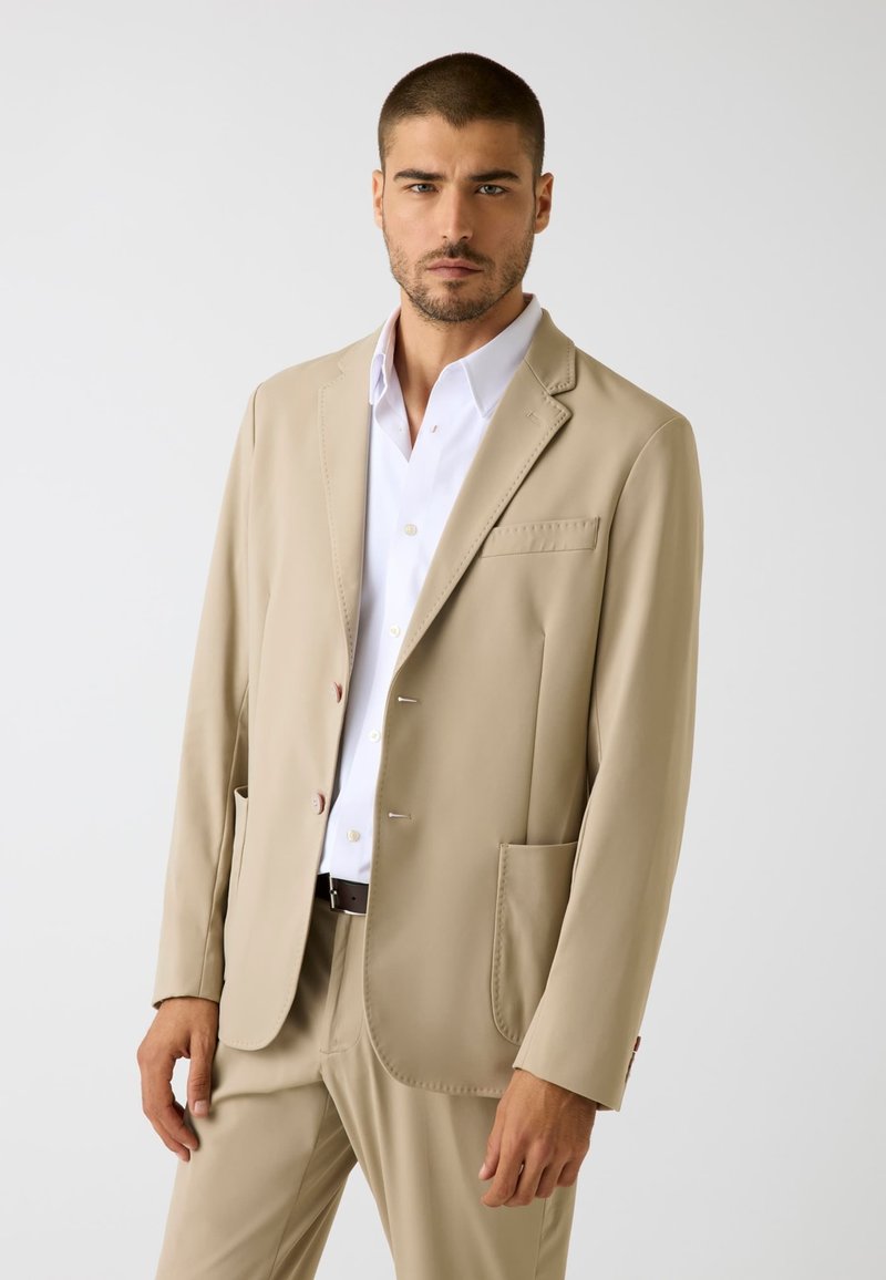 Giacca da completo beige realizzata in tessuto liscio, con rever a lancia, tre bottoni e due tasche frontali, abbinata a una camicia bianca.