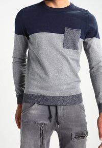 Pull en tricot bleu marine et gris avec un design à fente horizontale, poche poitrine texturée et poignets et ourlet côtelés. Associé à un pantalon cargo gris.