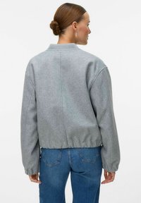 Femme avec un chignon portant une veste grise à manches longues et un jean bleu, vue de dos sur un fond uni.