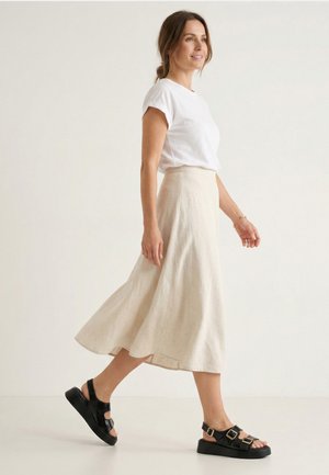 Frau geht drinnen, trägt ein weißes kurzärmeliges Hemd, einen beigen Midirock und schwarze Plateausandalen mit Schnallenriemen.