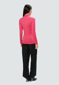 Figurbetontes, pinkes Langarmoberteil mit hohem Kragen, kombiniert mit schwarzen weiten Hosen und hohen Schuhen, mit einer glatten Textur.