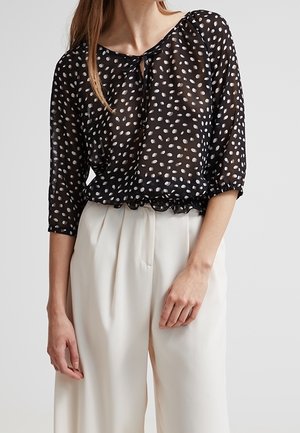 Frau trägt eine transparente schwarze Bluse mit weißen, unregelmäßigen Punkten und Dreiviertelärmeln, kombiniert mit weißen, hoch taillierten, weiten Hosen.