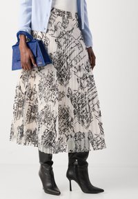 Vrouw draagt een geplooide witte rok met een zwart schetspatroon, zwarte haklaarzen, een lichtblauwe cardigan en houdt een blauw textuurhandtas vast.