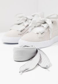 Paire de baskets beiges avec des lacets larges en satin, avec un jeu supplémentaire de lacets blancs texturés enroulés devant sur une surface blanche.