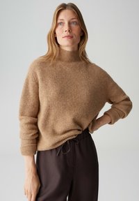 Pull beige côtelé avec un col montant et des manches retroussées, associé à un pantalon marron foncé à cordon. Texture lisse, coupe décontractée.