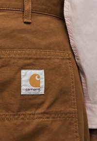 Τσέπη από καφέ παντελόνι εργασίας Carhartt με ραμμένη ετικέτα λογότυπου και μέρος από μανίκι ροζ πουκάμισου ορατό στη δεξιά πλευρά.