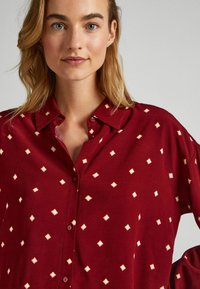Camisa burdeos con cuello abotonado, que presenta un patrón de rombos en crema, confeccionada en una tela suave con un corte ligeramente holgado.