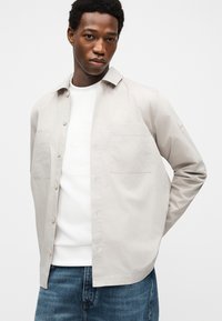 Νεαρός με κοντά σγουρά μαλλιά που φοράει ένα ανοιχτό μπεζ πουκάμισο πάνω από λευκό μπλουζάκι και μπλε τζιν, ποζάροντας μπροστά σε λευκό φόντο.