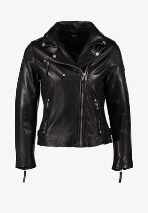 Femme marchant, portant une veste en cuir noire, un haut blanc, un jean skinny bleu, des bottines noires, tenant une pochette noire dans la main gauche.