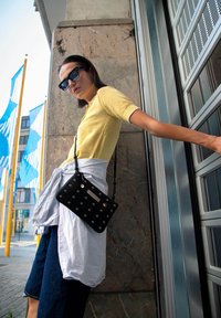 Zwarte crossbody tas met metalen oogdetails en een gestructureerde afwerking, gedragen over een lichtblauw shirt geknoopt in de taille. Gele top, donkere jeans.