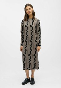 Ilga, smėlio spalvos megzta suknelė su juodais zigzago raštais, apvaliu iškirpimu ir ilgomis rankovėmis. Su juodais batais, kad kūrinys būtų užbaigtas.