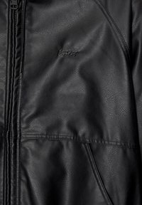 Primo piano di una giacca in pelle nera con zip visibile, cuciture stampate e un logo del marchio sobrio in rilievo su materiale testurizzato.