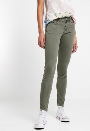Personne portant un jean skinny vert olive, des baskets montantes blanches et un haut léger à motif floral, debout devant un fond blanc.