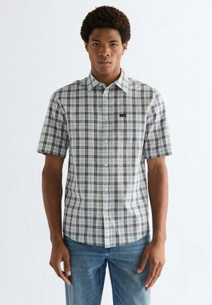 Uomo che indossa una camicia a quadri a maniche corte con bottoni sul davanti e jeans blu, in piedi contro uno sfondo bianco semplice.