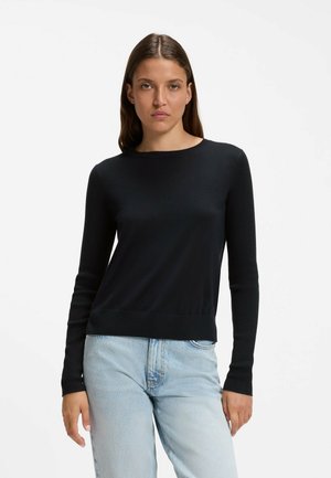 Frau mit langen braunen Haaren, die ein schwarzes langärmliges Shirt und hellblaue Jeans trägt, steht vor einem einfachen weißen Hintergrund.