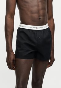 Άνδρας κορμός και πάνω μέρος των ποδιών με μαύρα μπόξερ Tommy Hilfiger, με λευκή ζώνη μέσης που φέρει το λογότυπο της μάρκας.