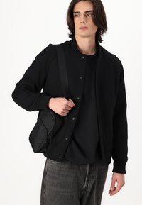 Svart sweatshirt med knäppning framtill, ribbade muddarna och en avslappnad passform. Modellen bär en liten svart axelväska.