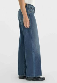 Jean en denim à jambes larges dans un délavage bleu foncé avec une texture lisse, présentant une poche avant et des détails de coutures contrastées.