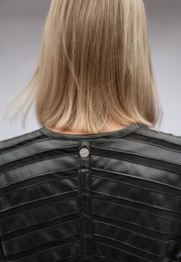 Blonde vrouw met steil haar tot op de schouders, gekleed in een zwarte leren jas met horizontale gestikte strepen, gezien van achteren.