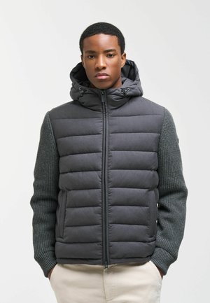 Junger Mann trägt eine dunkelgraue gesteppte Jacke mit gestrickten Ärmeln und Kapuze, steht mit den Händen in den Taschen einer hellbeigen Hose.