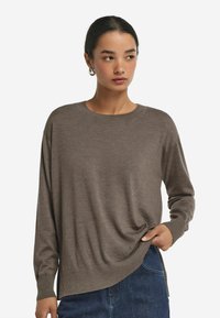 Brązowy sweter z długim rękawem, wykonany z miękkiego dzianiny, z okrągłym dekoltem i bocznymi rozporkami, zestawiony z ciemnoniebieskimi dżinsami.