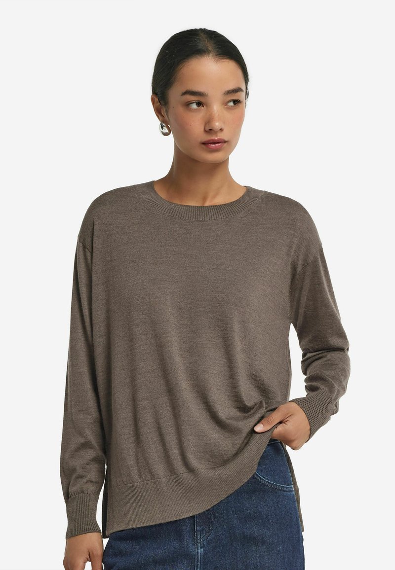 Brązowy sweter z długim rękawem, wykonany z miękkiego dzianiny, z okrągłym dekoltem i bocznymi rozporkami, zestawiony z ciemnoniebieskimi dżinsami.