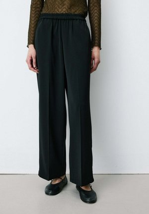 Personne portant un pantalon noir large et ample avec une ceinture élastique et des ballerines noires, debout contre un mur blanc.