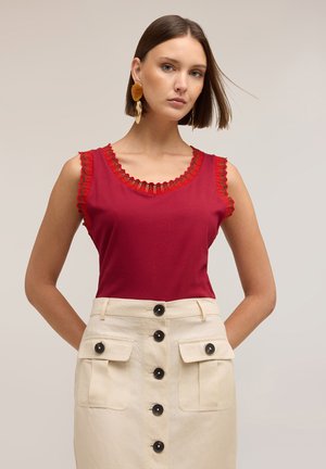 Frau mit glattem braunem Haar trägt ein ärmelloses rotes Oberteil mit Spitzenbesatz und einen hoch taillierten cremefarbenen Rock mit schwarzen Knöpfen und Fronttaschen.