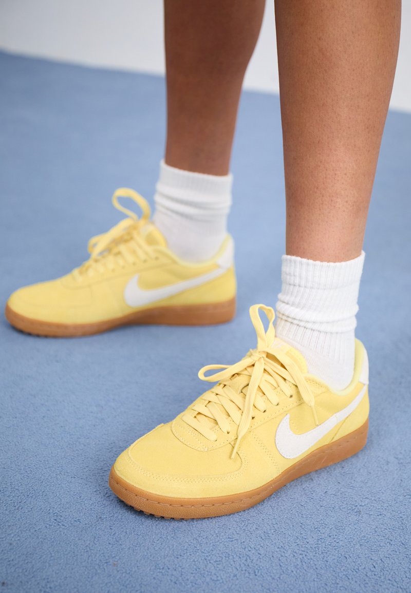 Zapatillas de ante amarillas con el logo de Nike en blanco y suela de goma, con cordones, textura suave, y llevadas con calcetas blancas.