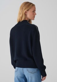 Pull en tricot bleu marine avec un col montant, des poignets et un ourlet côtelés. Présente des rayures blanches sur les côtés des manches. Coupe décontractée et oversize.