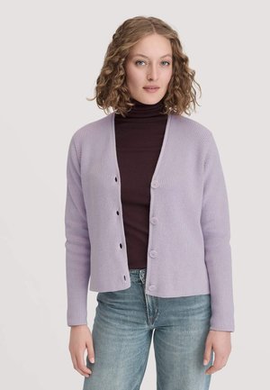 Frau mit lockigem Haar, die einen helllila Knopf-Cardigan über einem dunklen Rollkragenpullover und blauen Jeans trägt, und vor einem schlichten Hintergrund steht.