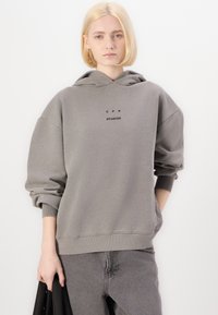 Hanorac gri cu glugă, având umeri lăsați, manșete ribbordate și un mic logo text negru "CPH STUDIOS" pe față.