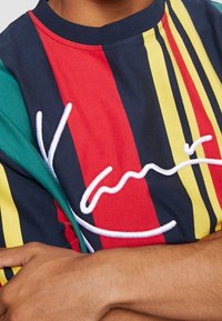 Flerfärgad kortärmad skjorta med djärva vertikala ränder i röd, blå, gul och grön; har vit broderad text.