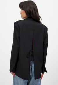 Schwarzer Blazer mit auffälligem Cutout auf der Rückseite, gebundenem Detail, strukturierten Schultern und langen Ärmeln mit seitlichen Schlitzen. Glatte Stofftextur.