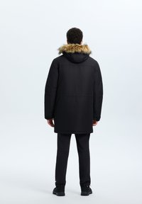 Parka nera con cappuccio foderato in pelliccia, con un design a taglio dritto, maniche lunghe e tessuto testurizzato, abbinata a pantaloni e scarpe nere.