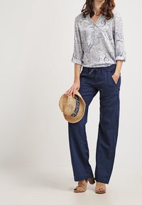 Blusa azzurra con motivo paisley e maniche arrotolabili, abbinata a pantaloni a gamba larga blu navy. Dettagli aggiuntivi includono un cappello di paglia e sandali nudi.