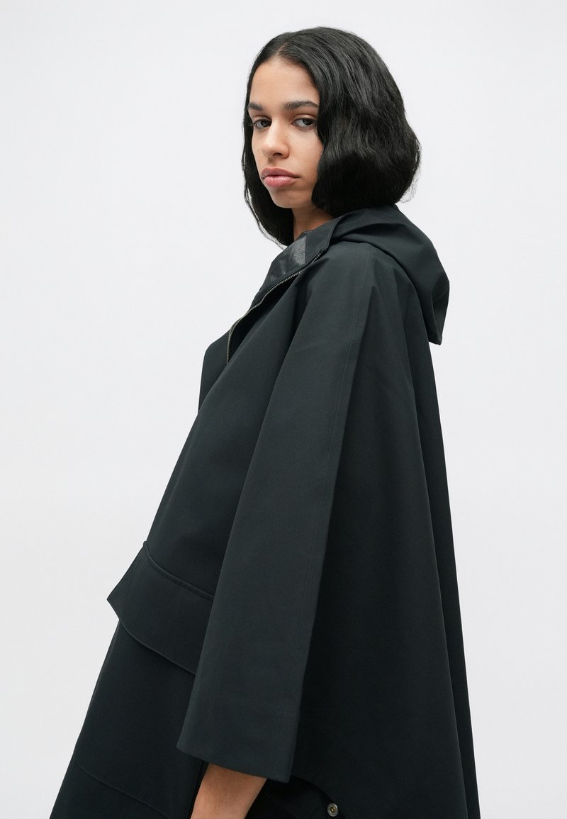 Black hooded cape with a loose fit, featuring a layered design, smooth texture, and front pocket detailing. Plain background.