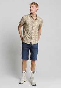 Beige shirt met korte mouwen en een schelpenpatroon, gecombineerd met denim shorts. Gemaakt met witte sokken met zwarte strepen en grijze sneakers.