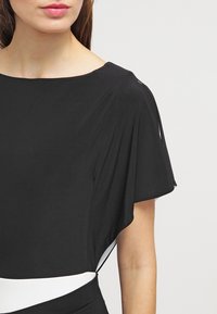 Svart klänning med en figursydd silhuett, rund halsringning och korta fällande ärmar. Har en vit accent vid midjan och en slät tygstruktur.