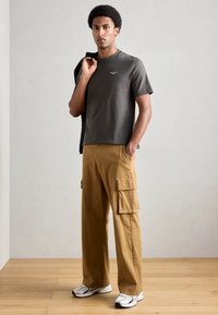 Pelēks īsām piedurknēm t-krekls ar mazu logo, saskaņots ar brūniem kargo šortiem, kas aprīkoti ar sānu kabatām un brīvu piegriezumu, pabeigts ar baltām sporta apaviem.