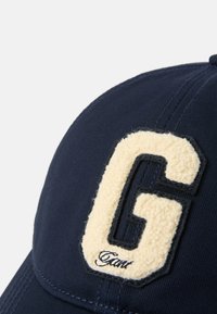 Gorra azul marino con un parche de letra "G" beige y suave, costuras detalladas y una textura blanda. Incluye el logo de la marca en cursiva debajo del parche.
