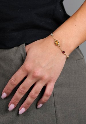 Main reposant sur la poche d'un pantalon gris, portant un bracelet chaîne en or avec des pierres précieuses ambre, rouges et claires, et un vernis à ongles rose clair.