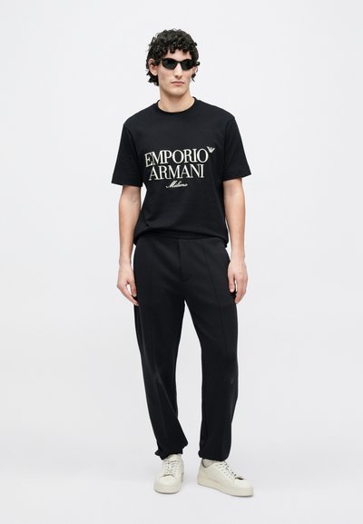 Μαύρο κοντομάνικο μπλουζάκι με λευκό λογότυπο "Emporio Armani Milano", σε συνδυασμό με μαύρο παντελόνι κομμένο στα μέτρα και ανοιχτό μπεζ αθλητικά παπούτσια. Μινιμαλιστικός σχεδιασμός.