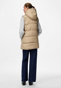 Beige Steppweste mit Kapuze, gestepptem Design und längerer Passform, kombiniert mit grauen Ärmeln und dunkelblauen weit geschnittenen Jeans. Schwarze Stiefel runden den Look ab.