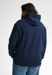 Hanorac bleu marin cu o glugă mare, ajustabilă. Prezintă o textură netedă și manșete și tiv nervurat. Vedere din spate cu croială lejeră.