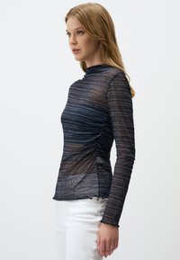 Maglietta a maniche lunghe in rete blu navy con strisce orizzontali argentate, collo alto, dettagli arricciati ai lati e orlo ondulato, abbinata a pantaloni bianchi.
