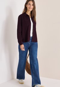 Maroon button-up skjorte, hvid t-shirt under, brede blå jeans og lyse sneakers. Brun håndtaske hængende over skulderen.