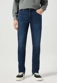 Donkerblauwe spijkerbroeken met een rechte snit, voorzien van voorzakken en een klassiek vijfzakkenontwerp. Draagt met zwart-witte sneakers.