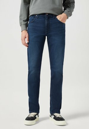 Jeans in denim blu scuro con taglio dritto, dotati di tasche frontali e un classico design a cinque tasche. Indossati con scarpe da ginnastica nere e bianche.